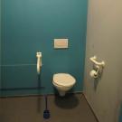 Het aangepast toilet heeft een vaste beugel aan de wand en een opklapbare beugel aan de open zijde van het toilet. De achterwand contrasteert goed met de witte toestellen. (Jeugdcentrum Ahoy, Wijnegem)